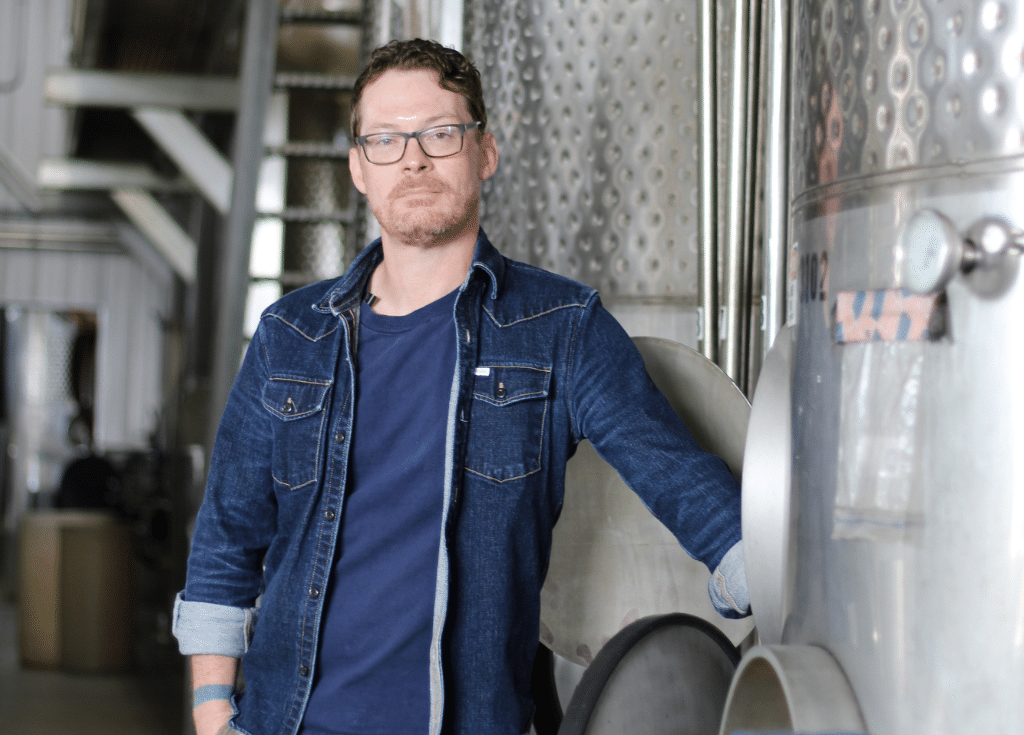 Winemaker Michael Reidy posing in front of a stainless steel tank in the winery production area.